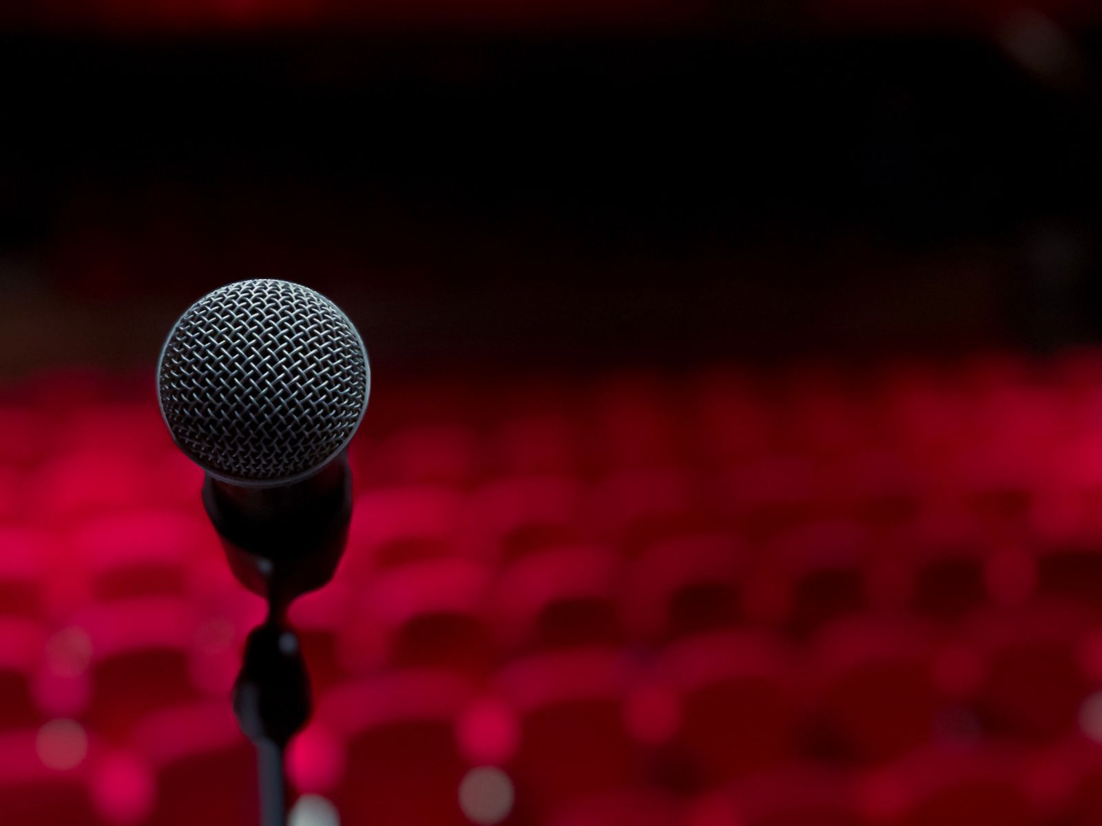 Microfone em pé sobre o palco, em close, com fileiras de poltronas vermelhas vazias desfocadas ao fundo, em ambiente escuro e dramaticamente iluminado