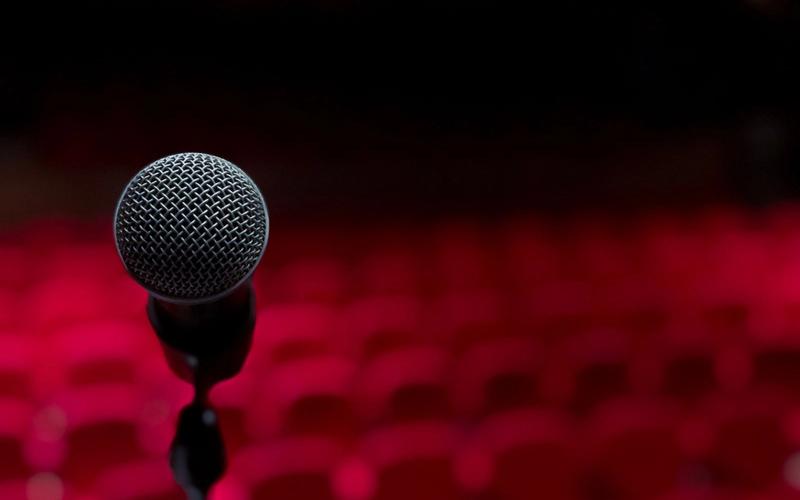 Microfone em pé sobre o palco, em close, com fileiras de poltronas vermelhas vazias desfocadas ao fundo, em ambiente escuro e dramaticamente iluminado