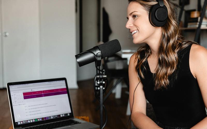 Mulher de fones sorrindo enquanto grava ao microfone num estúdio, com o software de edição de áudio aberto no notebook ao lado — os três pilares de um podcast: voz, estúdio e edição.