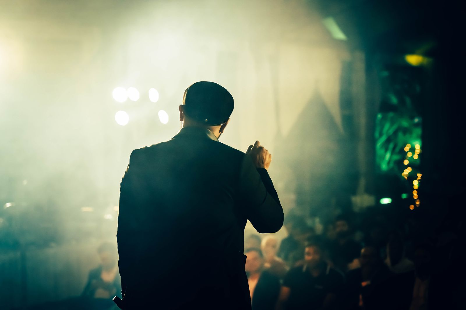 Pessoa de costas, em silhueta, falando diante de uma plateia em um ambiente com luz de palco intensa