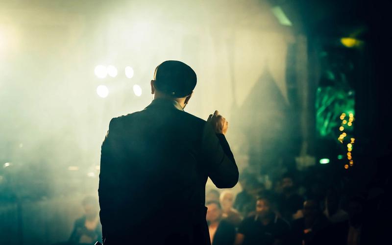 Pessoa de costas, em silhueta, falando diante de uma plateia em um ambiente com luz de palco intensa