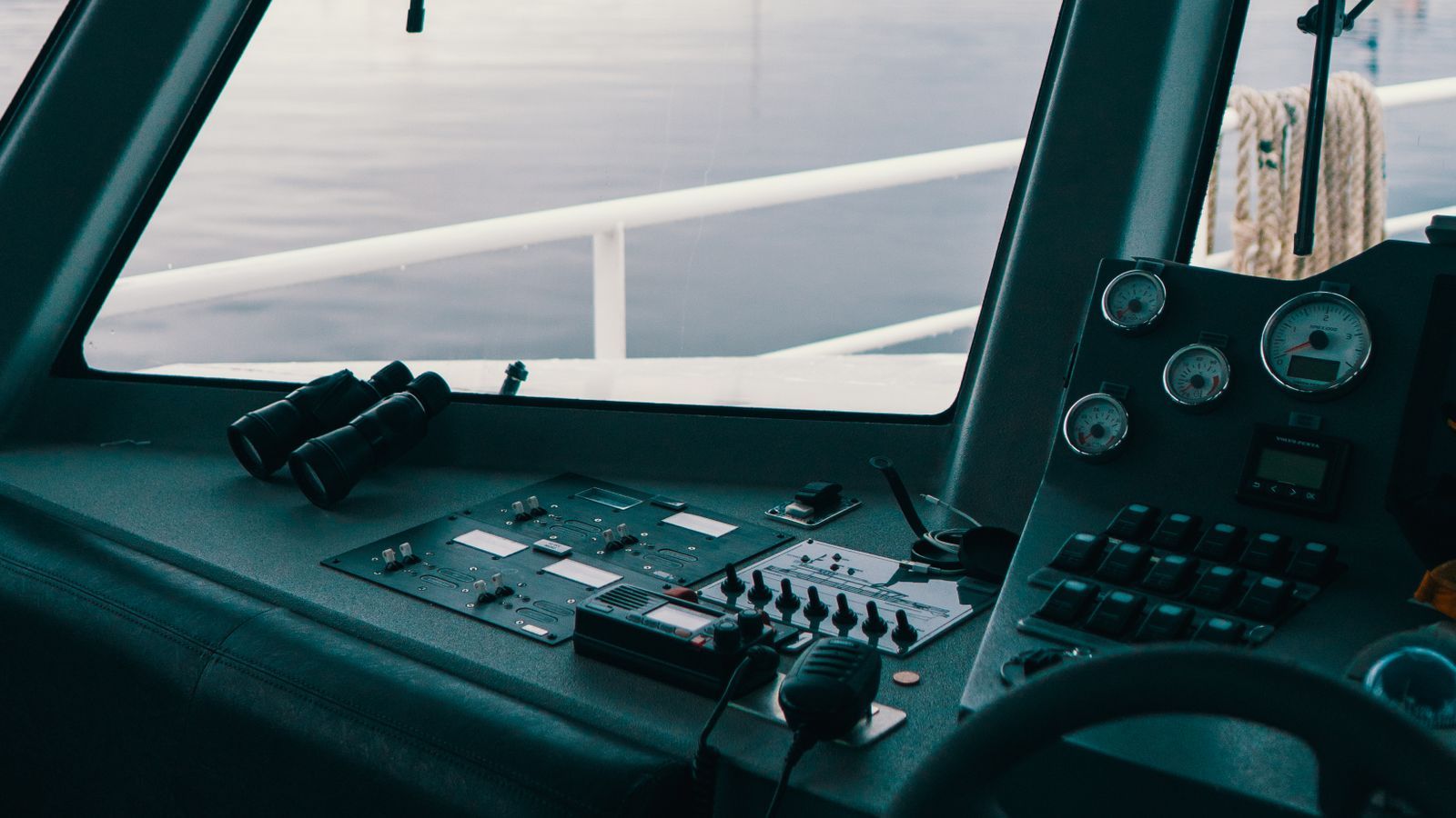 Console de comando de uma embarcação com rádio VHF marítimo, binóculos e instrumentos de navegação, com o mar ao fundo