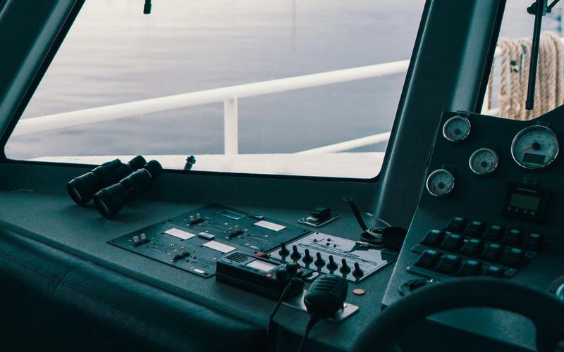 Console de comando de uma embarcação com rádio VHF marítimo, binóculos e instrumentos de navegação, com o mar ao fundo