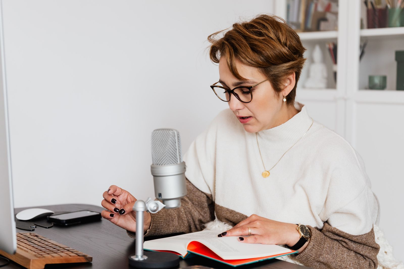 Mulher de óculos sentada à mesa, lendo em voz alta um livro aberto diante de um microfone de estúdio para gravar narração de audiolivro