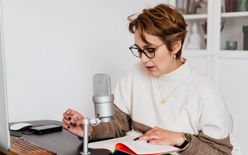Mulher de óculos sentada à mesa, lendo em voz alta um livro aberto diante de um microfone de estúdio para gravar narração de audiolivro