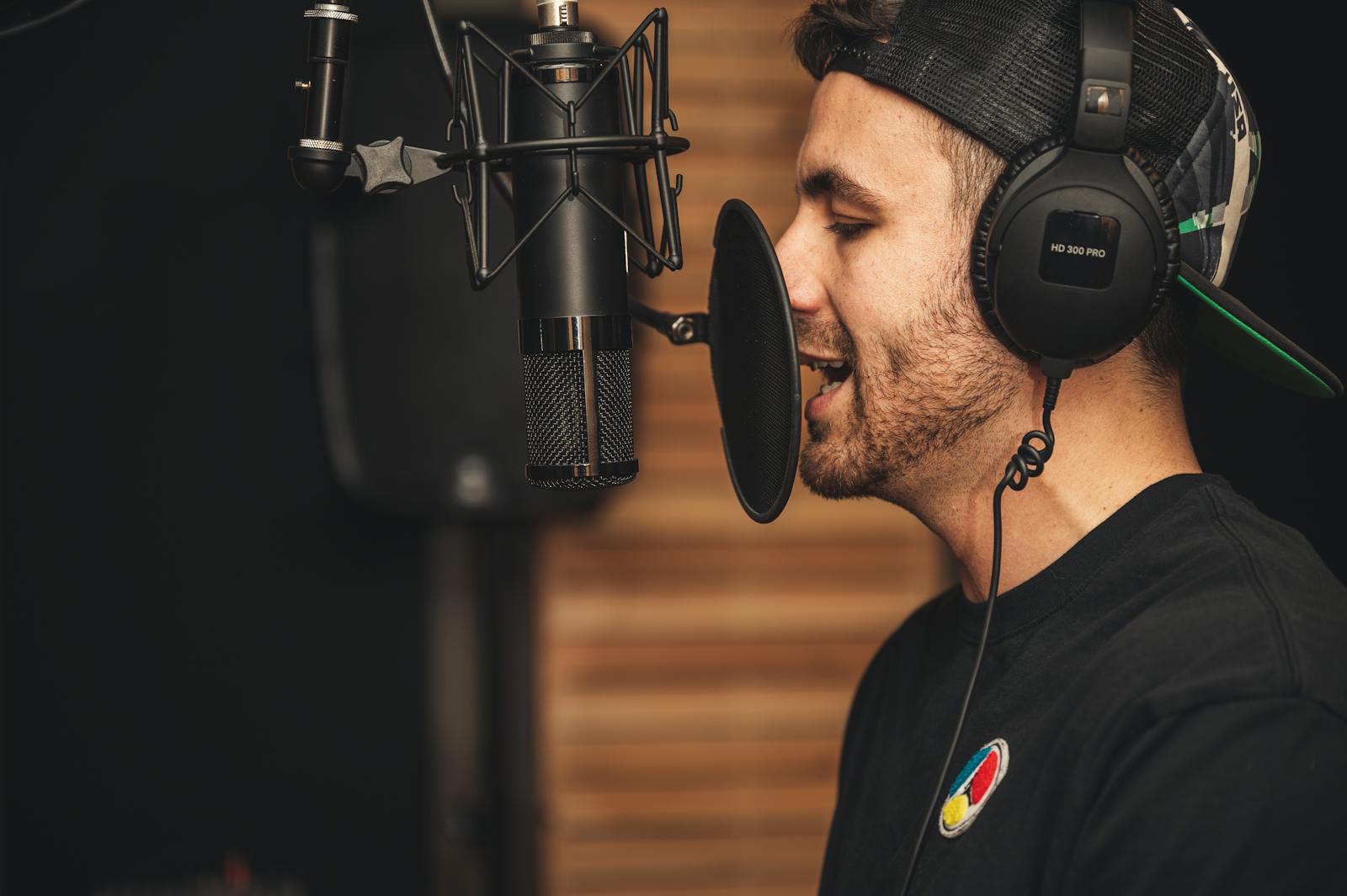 Homem de perfil, com fones de ouvido, interpretando um texto diante de um microfone profissional com filtro pop num estúdio de gravação, concentrado na entrega vocal.