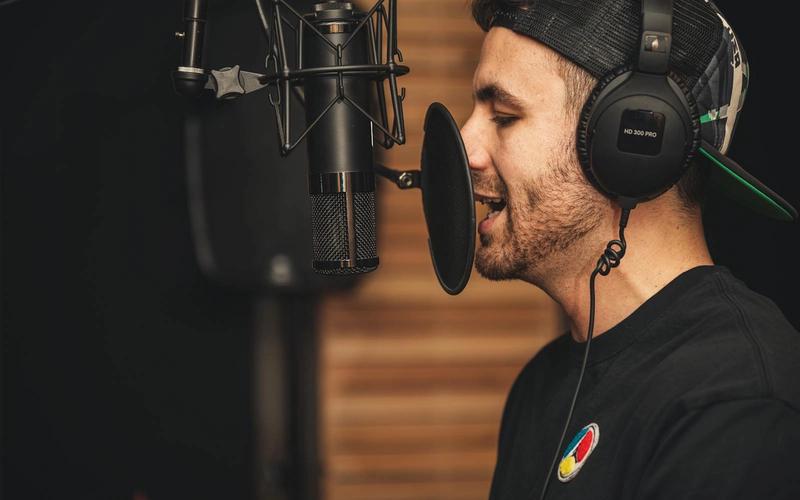 Homem de perfil, com fones de ouvido, interpretando um texto diante de um microfone profissional com filtro pop num estúdio de gravação, concentrado na entrega vocal.