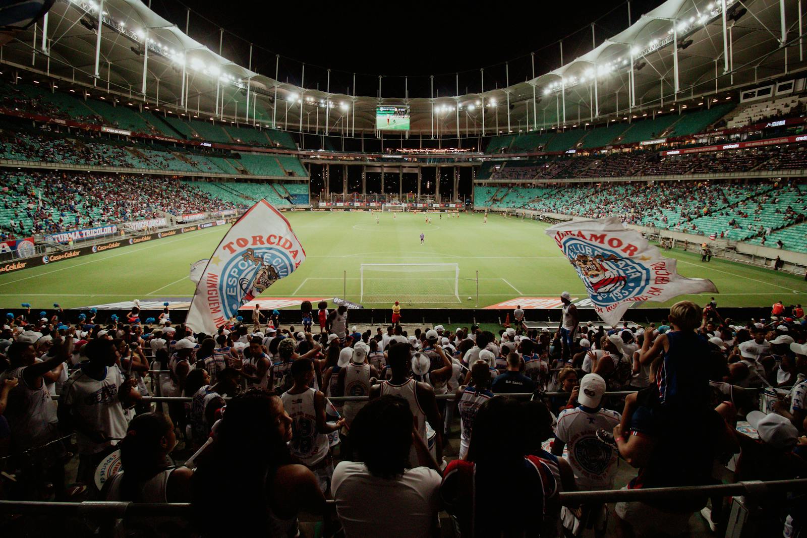 Estádio de futebol lotado à noite, visto da arquibancada: a torcida de costas agita grandes bandeirões enquanto o gramado verde brilha sob os refletores.