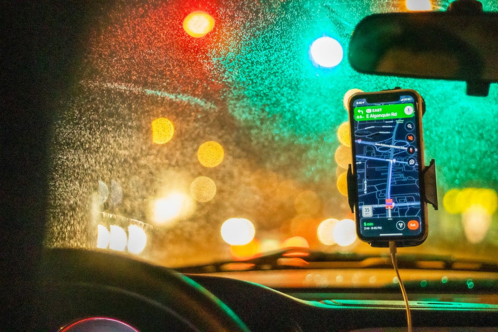 Celular com aplicativo de GPS preso ao painel do carro, mostrando instrução de rota à noite sob chuva, com luzes de trânsito desfocadas ao fundo