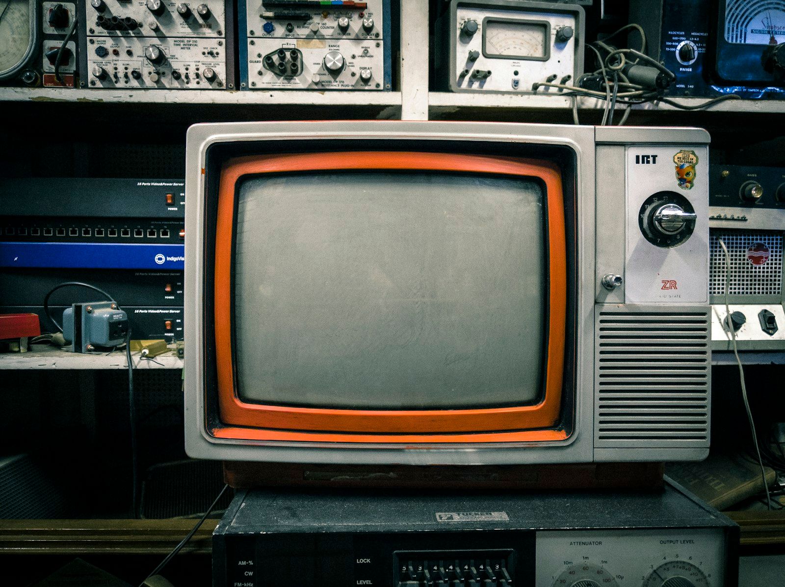 Televisão antiga de tubo (CRT), com moldura cinza e detalhe laranja e botão seletor de canais giratório, em cima de um armário, cercada por equipamentos eletrônicos vintage de rádio e transmissão em uma prateleira ao fundo, evocando a era clássica da televisão e do telejornalismo.