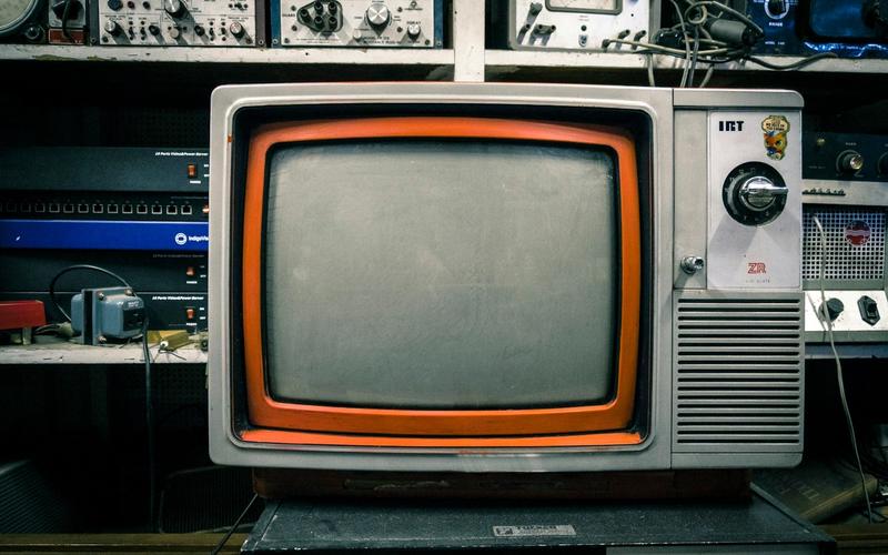 Televisão antiga de tubo (CRT), com moldura cinza e detalhe laranja e botão seletor de canais giratório, em cima de um armário, cercada por equipamentos eletrônicos vintage de rádio e transmissão em uma prateleira ao fundo, evocando a era clássica da televisão e do telejornalismo.