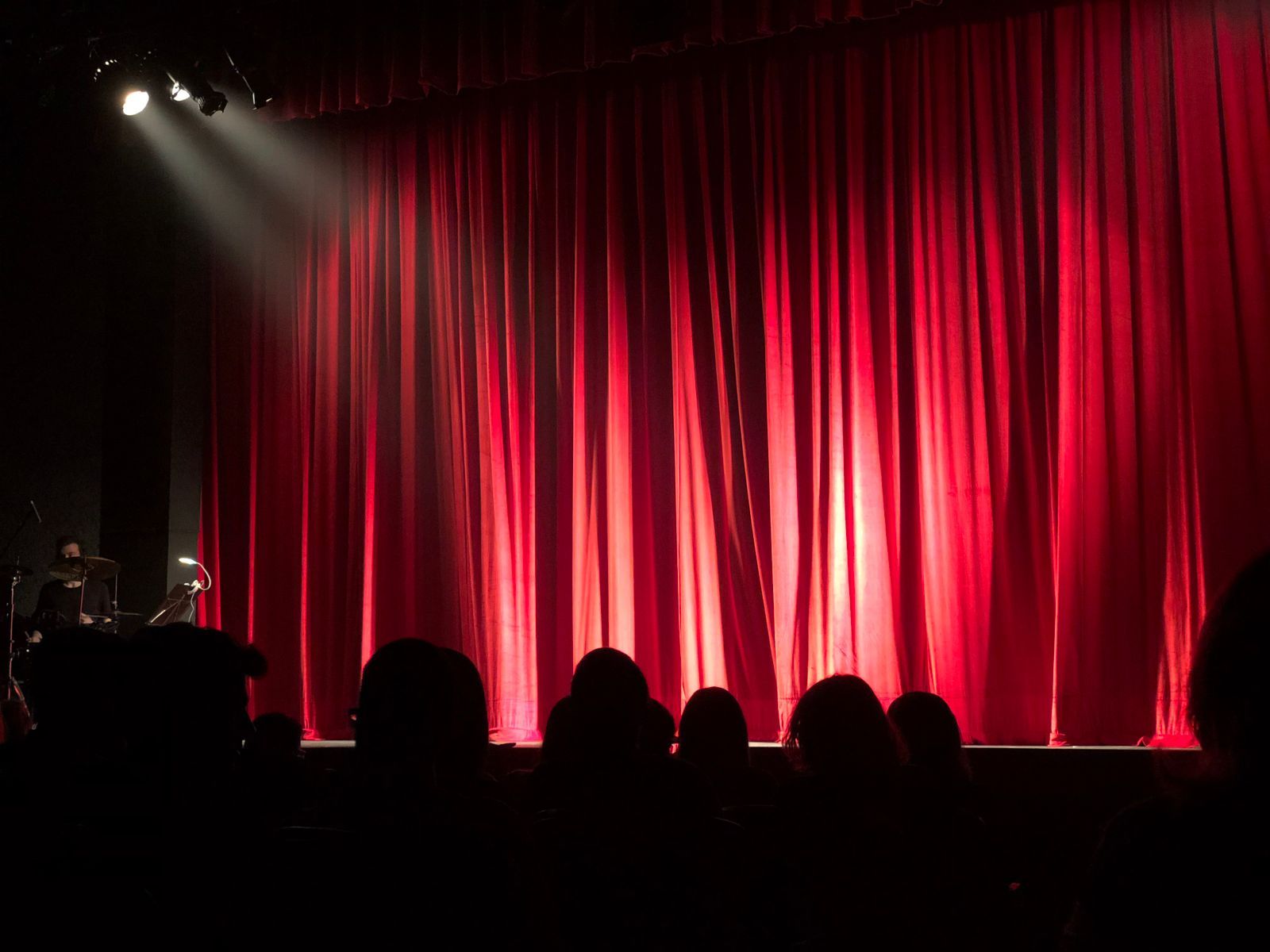 Palco de teatro com cortina vermelha iluminada por um spotlight, visto de trás das cabeças em silhueta da plateia durante uma apresentação ao vivo