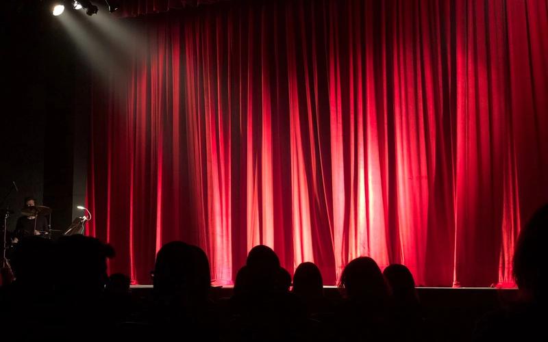 Palco de teatro com cortina vermelha iluminada por um spotlight, visto de trás das cabeças em silhueta da plateia durante uma apresentação ao vivo