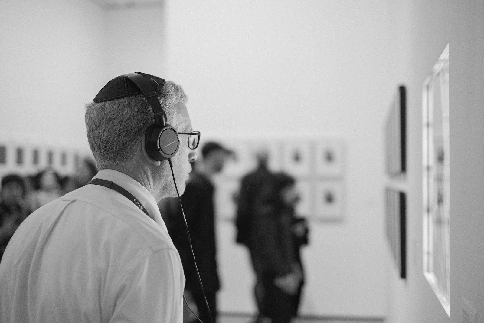 Visitante de museu usando fone de ouvido de audioguia, observando uma obra na parede de uma galeria, com outros visitantes desfocados ao fundo