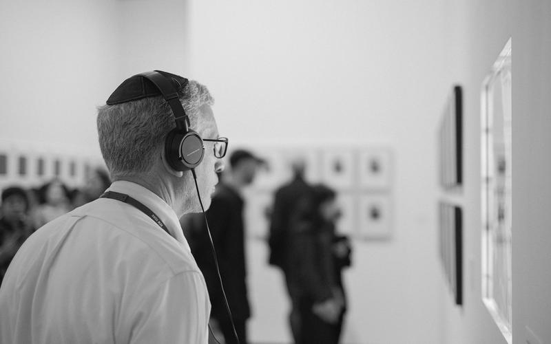 Visitante de museu usando fone de ouvido de audioguia, observando uma obra na parede de uma galeria, com outros visitantes desfocados ao fundo