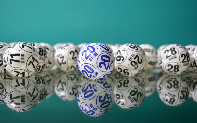 Fileira de bolinhas numeradas de sorteio, brancas com números em azul e preto, refletidas sobre superfície espelhada com fundo verde-azulado