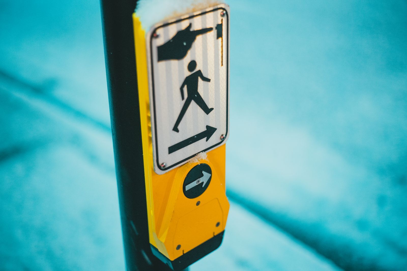 Close de um botão de travessia de pedestres acessível, com placa amarela mostrando ícone de pessoa atravessando e seta indicando o sentido, instalado em poste na rua