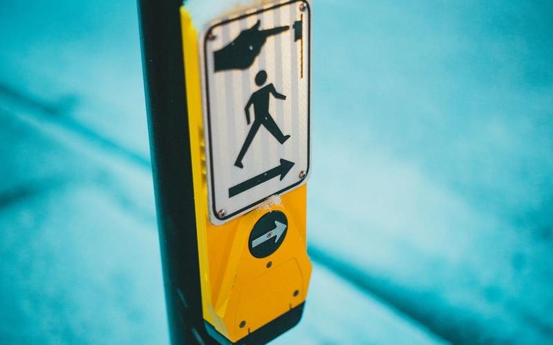 Close de um botão de travessia de pedestres acessível, com placa amarela mostrando ícone de pessoa atravessando e seta indicando o sentido, instalado em poste na rua