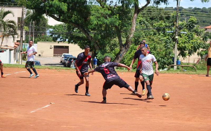 Jogadores disputam a bola durante uma partida de futebol de várzea em campo de terra, com casas e árvores ao fundo