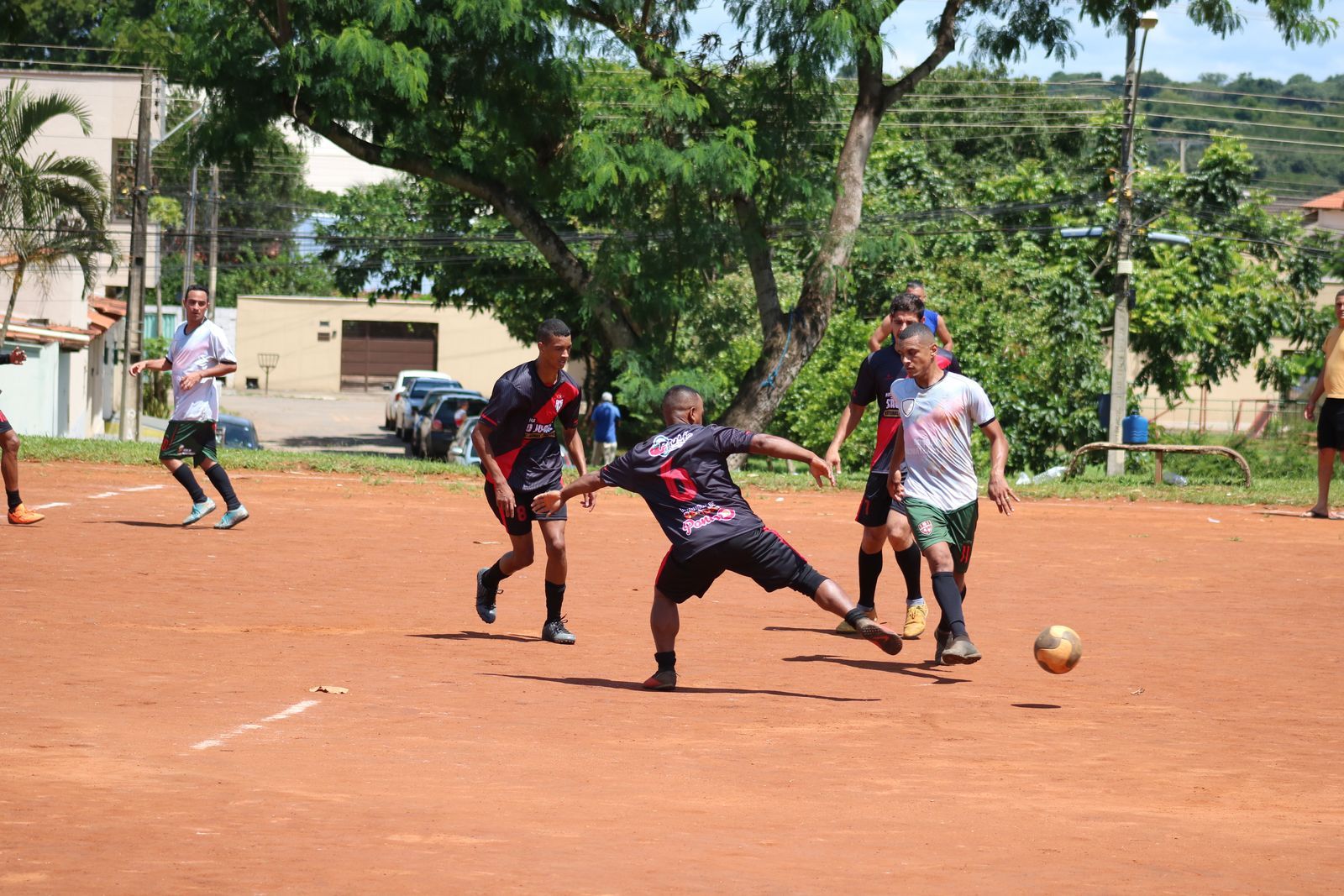 Jogadores disputam a bola durante uma partida de futebol de várzea em campo de terra, com casas e árvores ao fundo