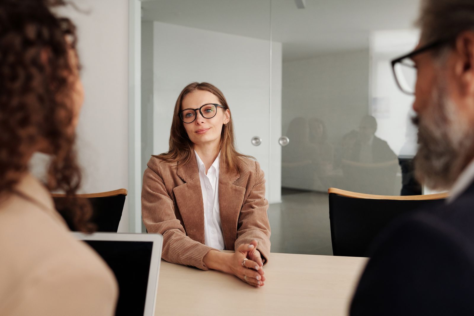 Candidata de blazer marrom sentada à mesa diante de dois entrevistadores durante uma entrevista de emprego em um escritório moderno e iluminado.