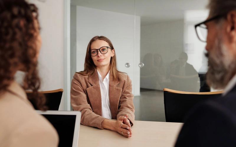Candidata de blazer marrom sentada à mesa diante de dois entrevistadores durante uma entrevista de emprego em um escritório moderno e iluminado.