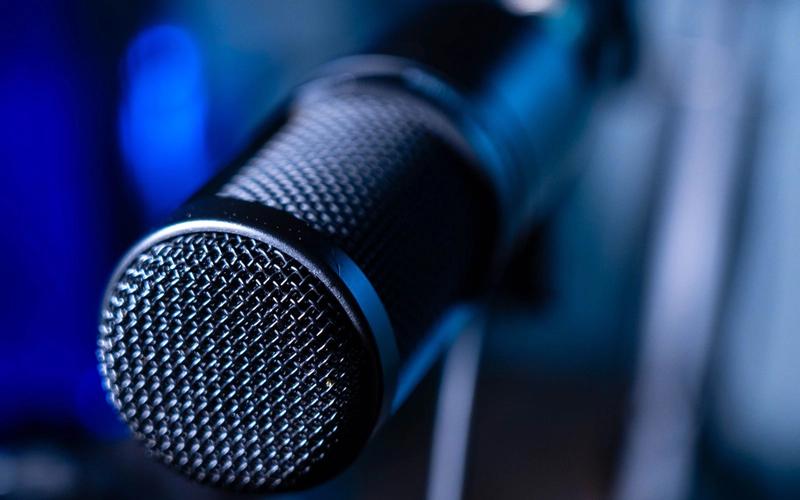Microfone profissional de estúdio em close, com fundo azul desfocado, evocando a locução e a transmissão de áudio do rádio indoor