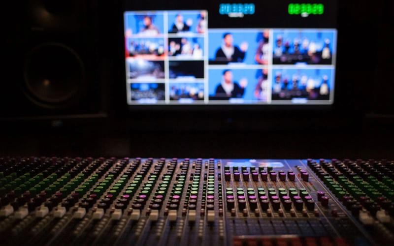 Mesa de mixagem profissional em primeiro plano, com tela ao fundo exibindo múltiplas câmeras em multivisão e contadores de tempo digitais, típica de uma sala de controle de transmissão ao vivo