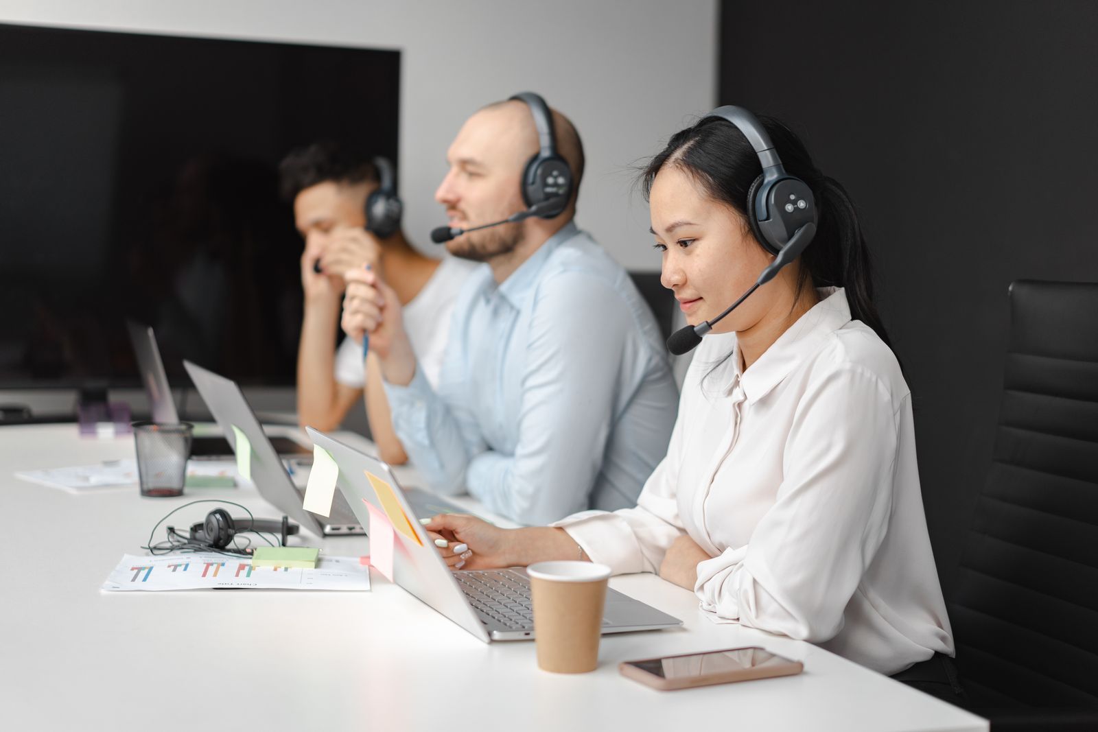 Três atendentes usando headsets trabalham lado a lado em notebooks, em uma central de atendimento telefônico moderna
