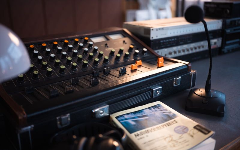Mesa de controle de um estúdio de rádio antigo, com mesa de som vintage cheia de botões coloridos, um microfone de bancada e equipamentos empilhados ao fundo