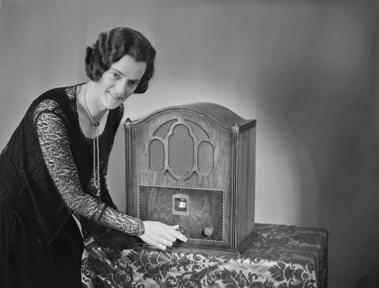 Fotografia em preto e branco de época: uma mulher com penteado e vestido dos anos 1930 ajusta o botão de um rádio de madeira sobre a mesa, evocando a era de ouro do rádio.