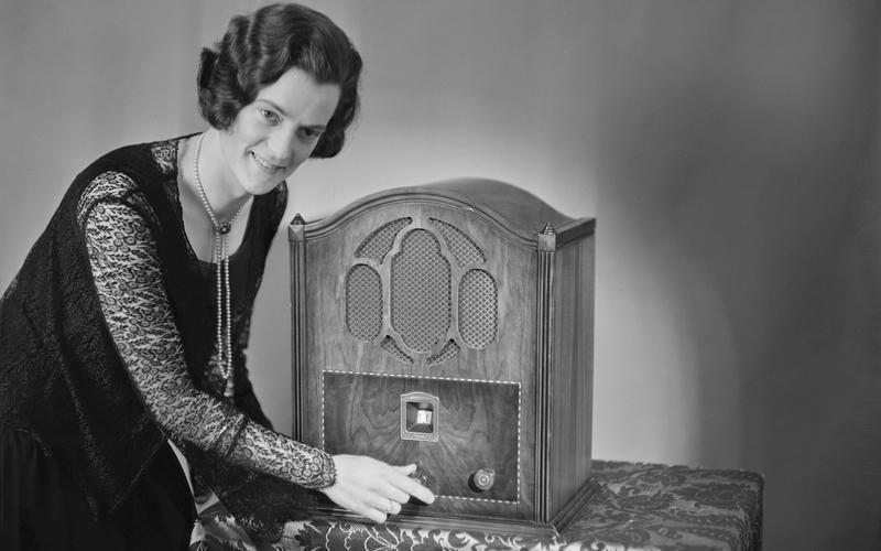 Fotografia em preto e branco de época: uma mulher com penteado e vestido dos anos 1930 ajusta o botão de um rádio de madeira sobre a mesa, evocando a era de ouro do rádio.