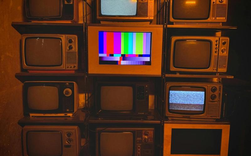 Parede com diversos televisores antigos empilhados, um deles ao centro exibindo as coloridas barras de teste de cor da TV.