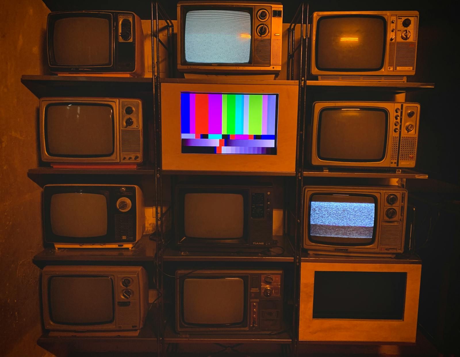 Parede com diversos televisores antigos empilhados, um deles ao centro exibindo as coloridas barras de teste de cor da TV.