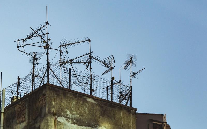 Emaranhado de antenas improvisadas e arame farpado no topo de um prédio antigo, contra um céu azul limpo, remetendo à estética de transmissões de rádio não oficiais