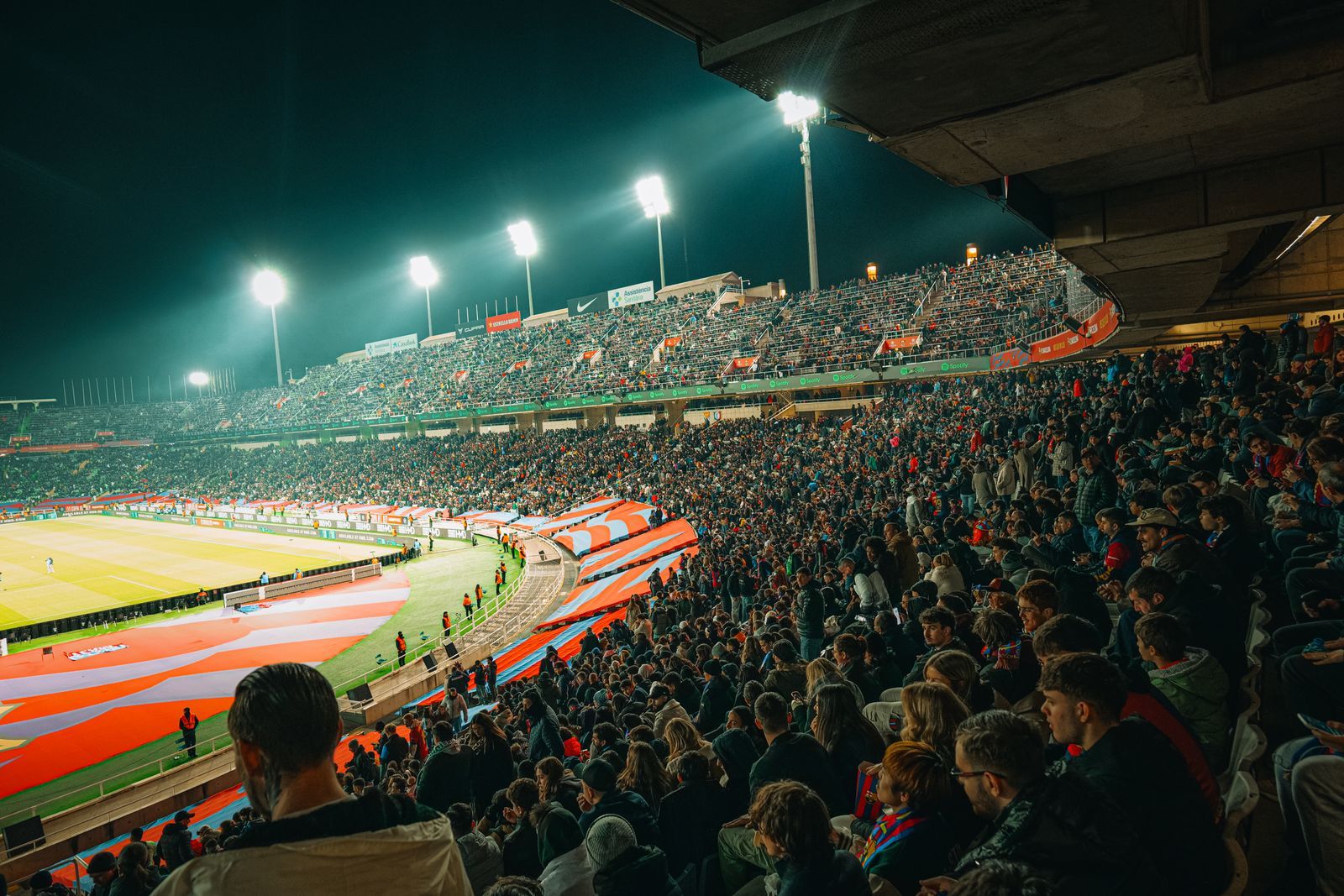 Estádio lotado à noite durante uma partida de futebol, com torcida animada sob os holofotes