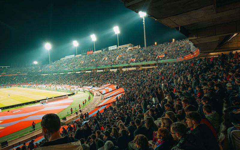 Estádio lotado à noite durante uma partida de futebol, com torcida animada sob os holofotes