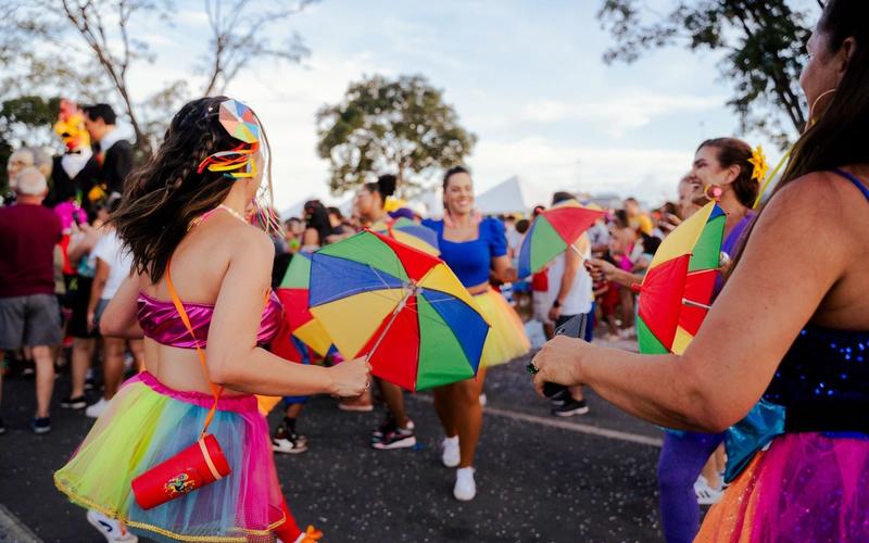 Foliãs dançam com sombrinhas coloridas em bloco de carnaval de rua no Brasil, cercadas por multidão animada ao entardecer