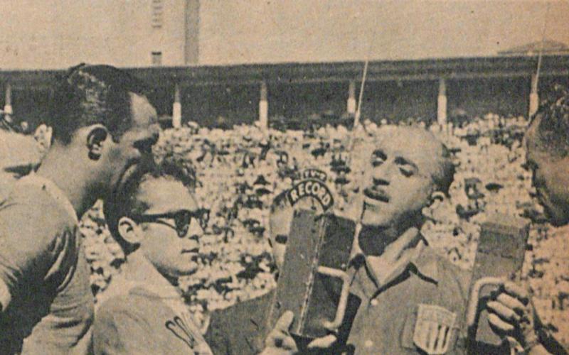 Foto em preto e branco de 1955 mostra o jovem jornalista Silvio Luiz, com óculos escuros sobre a cabeça, segurando um microfone da Rádio Record para entrevistar o árbitro Pedro Calil em um estádio de futebol lotado.