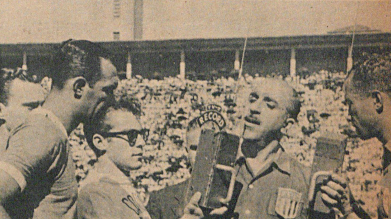 Foto em preto e branco de 1955 mostra o jovem jornalista Silvio Luiz, com óculos escuros sobre a cabeça, segurando um microfone da Rádio Record para entrevistar o árbitro Pedro Calil em um estádio de futebol lotado.