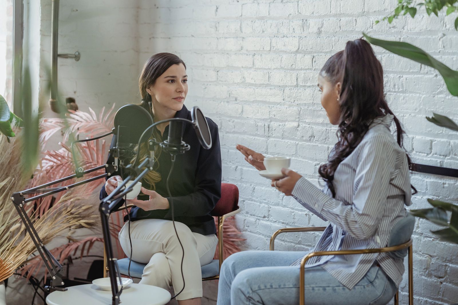 Entrevistadora ouvindo com atenção uma convidada durante uma entrevista, com microfone e xícara de café sobre a mesa em um estúdio iluminado