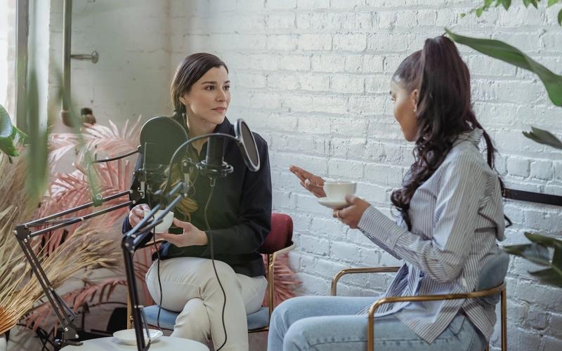 Entrevistadora ouvindo com atenção uma convidada durante uma entrevista, com microfone e xícara de café sobre a mesa em um estúdio iluminado