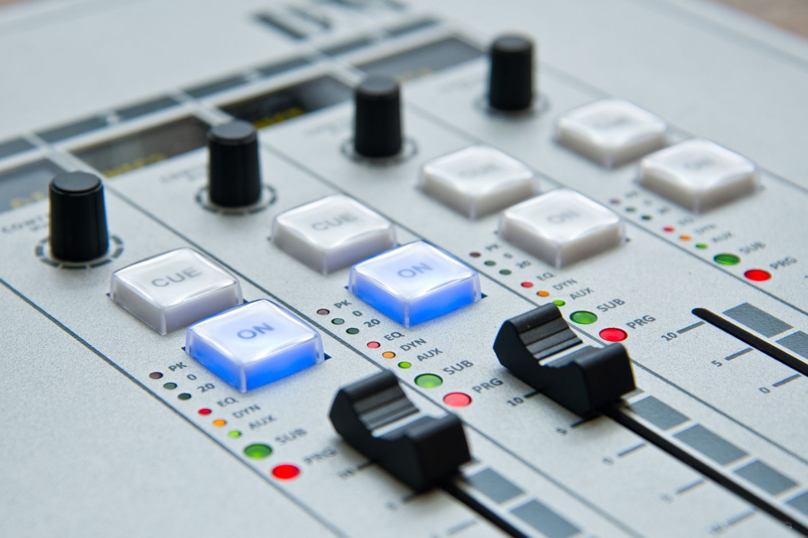 Close-up de painel de controle profissional de rádio, com botões ON e CUE iluminados em azul e faders de áudio, remetendo ao momento em que a emissora entra no ar para se identificar