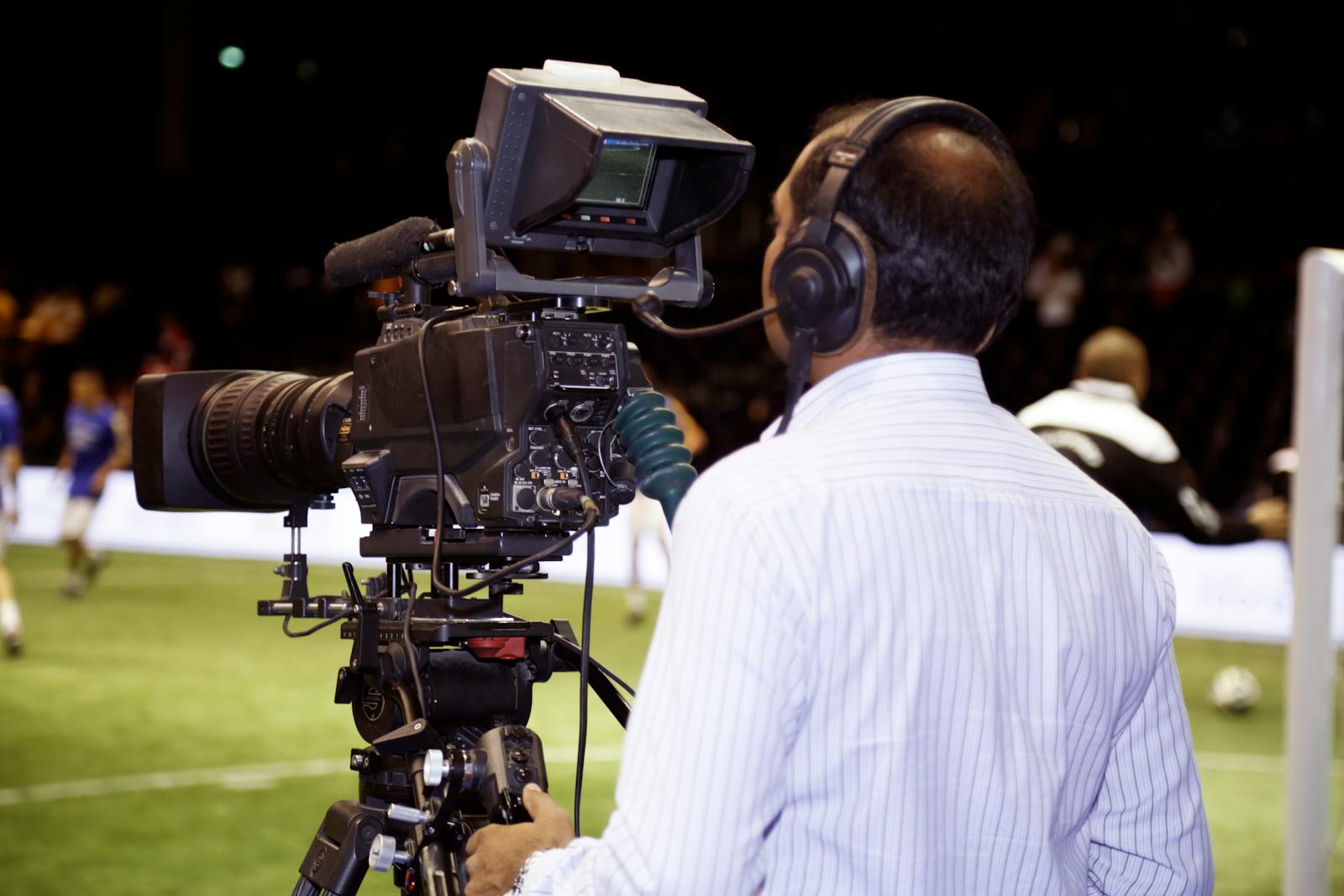 Profissional de transmissão esportiva com headset trabalhando à beira do gramado durante um jogo de futebol em um estádio.