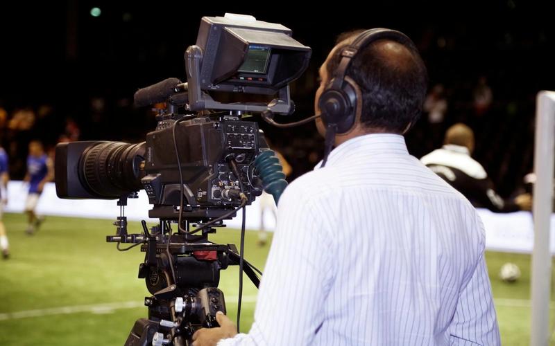 Profissional de transmissão esportiva com headset trabalhando à beira do gramado durante um jogo de futebol em um estádio.