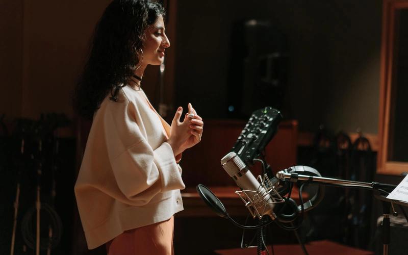 Mulher de perfil em um estúdio de gravação, em pé diante de um microfone condensador com pop filter, com as mãos juntas e expressão concentrada, como quem se prepara antes de usar a voz.