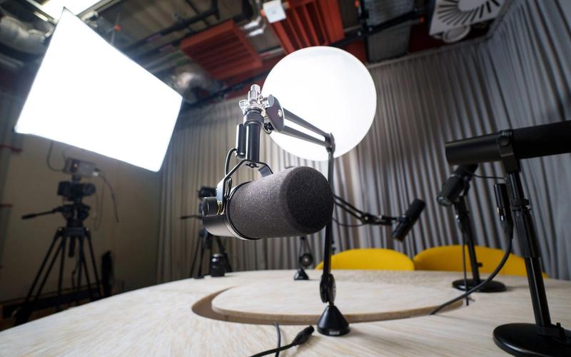 Estúdio de podcast moderno visto em grande angular: microfone profissional em primeiro plano, câmeras em tripé, luz softbox e cadeiras amarelas ao fundo, set pronto para gravação simultânea de áudio e vídeo