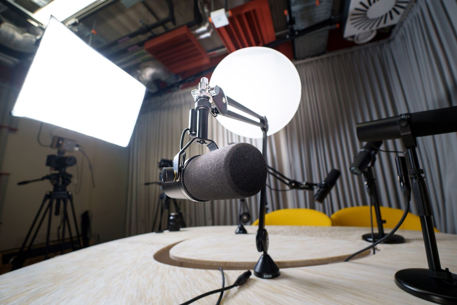 Estúdio de podcast moderno visto em grande angular: microfone profissional em primeiro plano, câmeras em tripé, luz softbox e cadeiras amarelas ao fundo, set pronto para gravação simultânea de áudio e vídeo