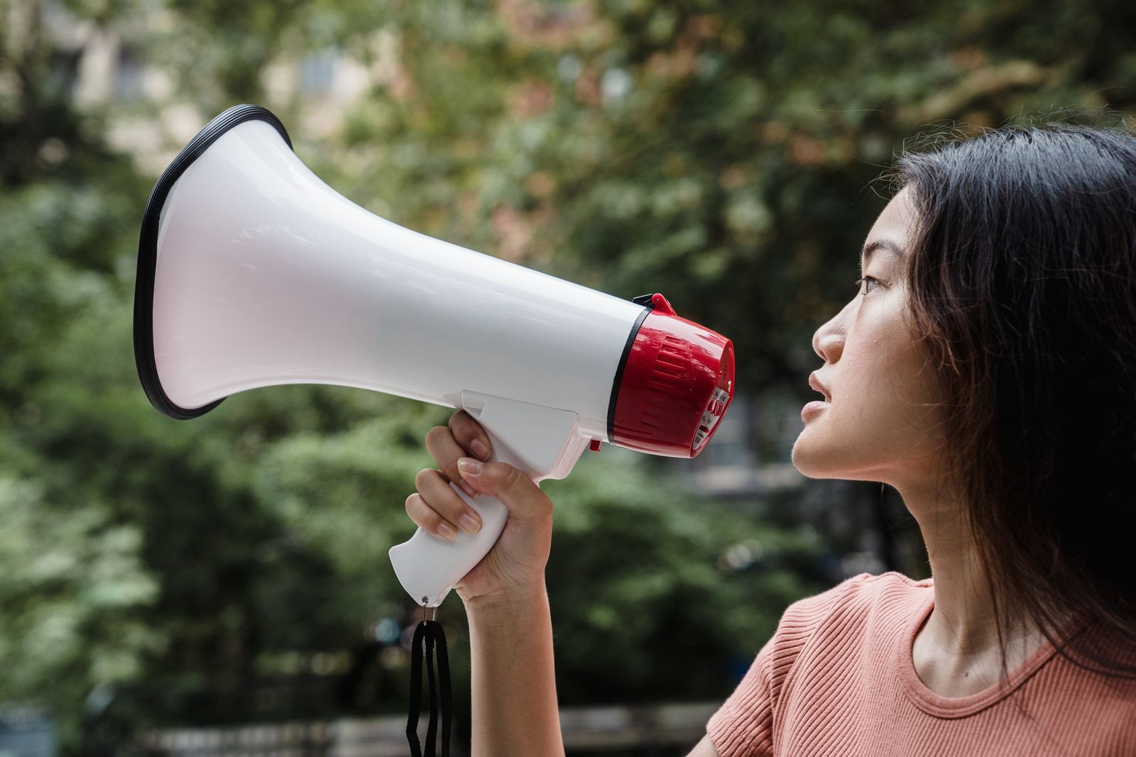Mulher jovem falando ao megafone, de perfil, em ambiente externo com árvores ao fundo, ilustrando o papel do tom de voz na comunicação.