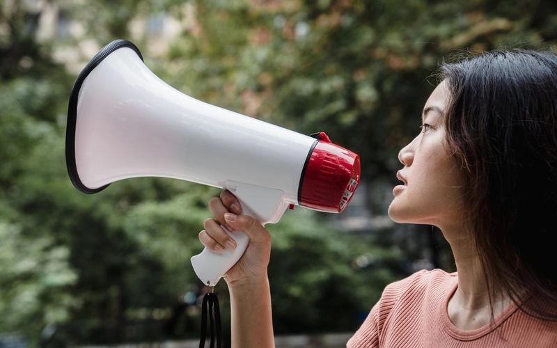 Mulher jovem falando ao megafone, de perfil, em ambiente externo com árvores ao fundo, ilustrando o papel do tom de voz na comunicação.